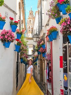 a woman in a yellow dress is walking down an alleyway with hanging flower pots