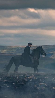 a man riding on the back of a black horse across a rocky field under a cloudy sky