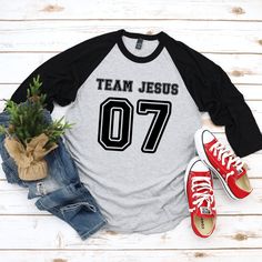 This Team Jesus Jersey Shirt is the perfect Christian shirt for any sports fan! It's a sporty stylish way to share your faith and heart for Jesus with anyone! Plus it's super soft and comfy. Whether your heading to your favorite sports game, running errands or a weekend get away, this cute Team Jesus shirt is a comfy casual Jesus shirt for any occasion. A stylish spin on the classic baseball raglan. The combed cotton blend makes it super soft, comfortable, and lightweight. You don’t even have to Casual Athletic Heather T-shirt For Game Day, Casual T-shirt With Lettering For Game Day, Varsity Sports Top With Lettering, Sports Fan Apparel Tops With Lettering, Casual T-shirt With Lettering For Sports Events, Casual Lettering T-shirt For Sports Events, Game Day Team-colored T-shirt With Lettering, Casual Game Day T-shirt With Lettering, Team-colored T-shirt With Lettering For Game Day