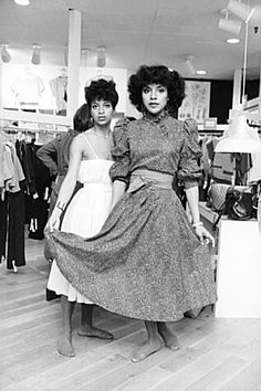 two women in dresses standing next to each other on a wooden floor with clothing racks behind them