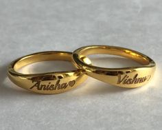 two gold wedding rings with names engraved on the sides and hearts in between them, sitting on a white surface