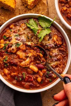 two bowls filled with chili and beans on top of a wooden table next to bread