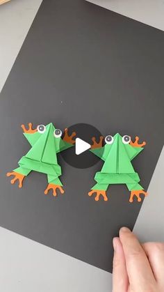 two paper frogs are facing each other on a black and gray surface with one frog looking at the camera