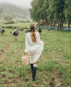 a woman in white dress walking through field with cows behind her and carrying a wicker basket