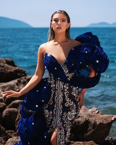 a woman in a blue dress standing on rocks near the ocean with her arms outstretched