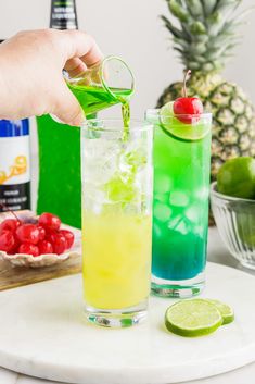 two glasses filled with different colored drinks next to pineapples and lime wedges