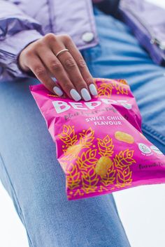 a woman with white nails holding a bag of chips in her hand and wearing blue jeans