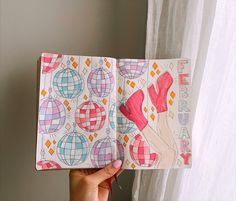 a hand holding an open notebook with colorful designs on the pages and in front of a window