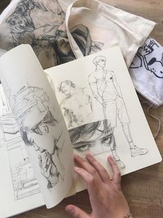 a person holding an open book on top of a wooden table next to some drawings