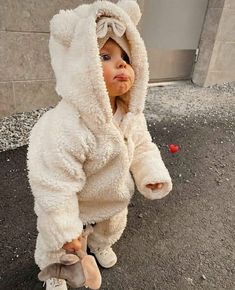 a toddler in a white teddy bear costume standing on the sidewalk with her hands behind her head