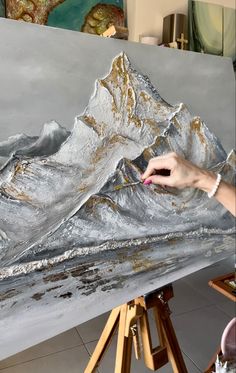 a person is painting a mountain scene with silver paint on an easel, while another hand holds a brush in the foreground