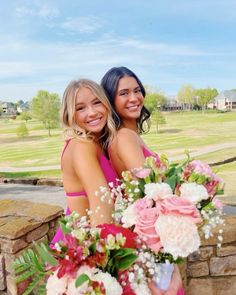 two beautiful young women standing next to each other in front of a stone wall holding flowers