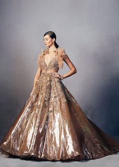 a woman in a gold gown with feathers on her head and shoulders, standing against a gray background