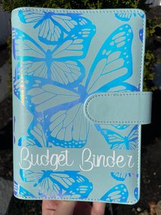 a hand holding a blue and white butterfly binder with the words budget binder on it