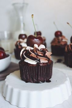 chocolate cupcakes with cherries and whipped cream are on a white cake plate