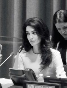 two women sitting at a table with microphones in front of them and one woman standing behind the podium