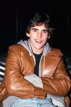 a young man wearing a brown leather jacket sitting on top of a metal bench with his arms crossed