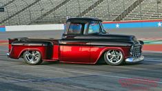 an old red and black truck driving on a track in front of some bleachers