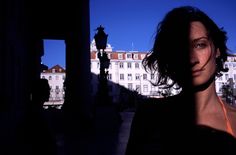 a woman standing in front of a building with her shadow on the ground and buildings behind her