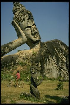 a man riding a bike past a large statue in the middle of a grass field