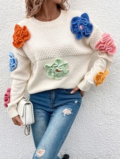 a woman wearing ripped jeans and a white sweater with colorful flowers on the sleeves is leaning against a wall