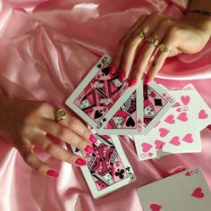 a woman's hands holding four playing cards on a pink satin surface with hearts