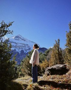 a woman standing on top of a lush green hillside next to a snow covered mountain