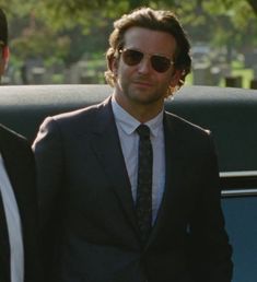 two men dressed in suits and sunglasses standing next to each other near a car with trees in the background