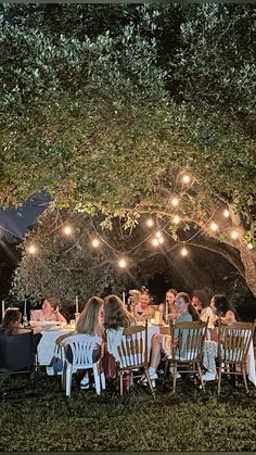 a group of people sitting around a table under a tree with lights strung from it