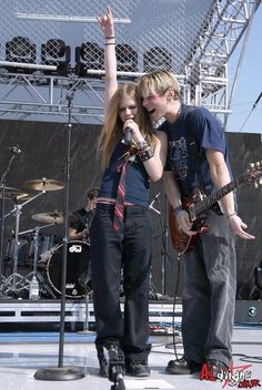 two people standing on stage with guitars and microphones in front of an outdoor concert