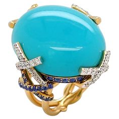 The polished blue-green beauty of turquoise is enhanced by precious gemstones of diamonds and sapphires for this ring. An oval cabochon turquoise is an apex, held in place by intercrossed 18k yellow gold strands with pavé-set brilliant cut diamonds. The basket features pavé-set sapphires. On the sides of the lustrous 18k yellow gold split shank is interlace of more sapphires. The total weight is 31.38 carats of an oval cabochon of turquoise measuring 21 by 25mm, 0.7 carats of 55 round brilliant Luxury Blue Turquoise Gemstone Ring, Luxury Turquoise Jewelry With Halo Setting, Kubler Ross, Round Sapphire, Bangles Jewelry Designs, Split Shank, Green Beauty, Bangles Jewelry, Precious Gemstones