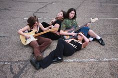 three young men sitting on the ground with guitars in their hands and one man holding an electric guitar