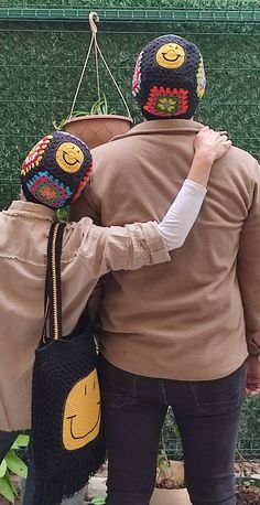 two people wearing knitted hats and holding onto each other's back while standing in front of a green fence