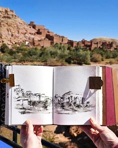 a person holding up an open book in front of a mountain range with houses on it