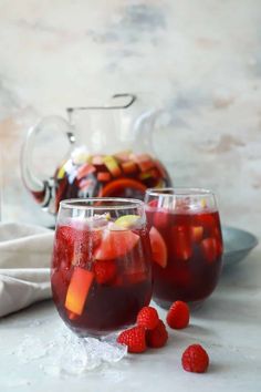 two glasses filled with red liquid and some raspberries on the table next to a pitcher