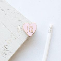 a pink heart shaped pin with the words you can on it next to a pencil