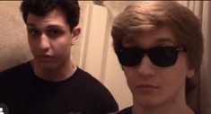 two young men standing next to each other in front of a door wearing black sunglasses