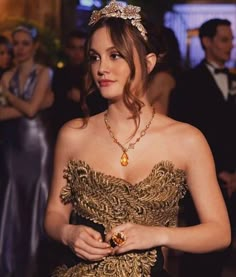 a woman wearing a gold dress with a tiara on her head and holding a piece of jewelry in her hand