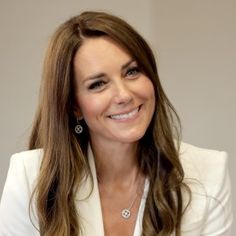 a woman with long brown hair wearing a white blazer and diamond necklace smiling at the camera