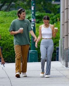 a man and woman walking down the street holding coffee cups in their hands while talking to each other