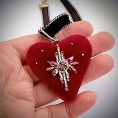 a hand holding a red heart shaped brooch with jewels on it's side