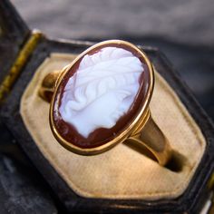 This storied ring showcases an oval hardstone cameo of a person wearing a headband, possibly laurel leaves. There are slight nicks on the hardstone, but nothing distracting. The ring is crafted in 10k yellow gold which we have left unpolished, and is currently a size 6.5. Luxury Vintage Cameo Signet Ring, Classic Oval Cameo Rings, Timeless Oval Intaglio Signet Ring, Timeless Oval Signet Ring With Intaglio, Formal Cameo Signet Ring, Classic Oval Cameo Jewelry, Heirloom Oval Cameo Signet Ring, Timeless Oval Intaglio Ring, Timeless Oval Ring With Intaglio Detail