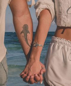 two people holding hands with tattoos on their arms near the ocean and sand in front of them