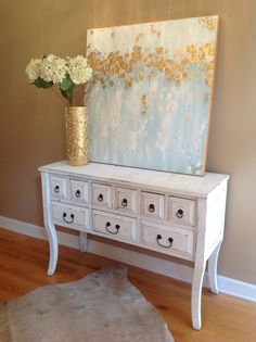 a white dresser with drawers and flowers on it in front of a large art piece