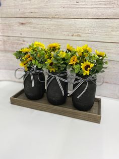 three black mason jars with sunflowers tied to them on a wooden tray in front of a white wall