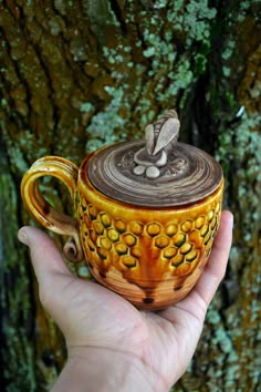 a hand holding a ceramic teapot in front of a mossy tree trunk with leaves on it