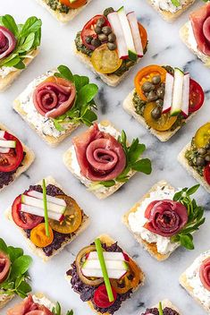 many small sandwiches with different toppings are arranged on a marble surface, including tomatoes and olives