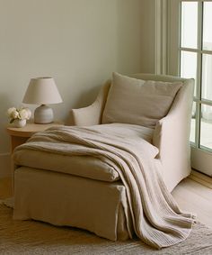 a white chair with a blanket on it next to a lamp and window sill