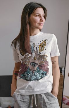 "CRYSTALLINE (UNISEX TEE) Designed and handmade in Phoenix. The witchy works! This hand-drawn design features a witchy hand with a third eye, holding a beautiful crystal, surrounded by flowers, mushrooms / fungi, and gorgeous butterflies. This designs is truly a unique showstopper. ★ABOUT OUR UNISEX TEES★ Our unisex tees are super soft, long and lean. Customers consistently mention the tees run large, so order down for a tighter fit. Our cream tees are 100% cotton; our white and certain colors a Mystical Cotton Tops With Graphic Print, Mystical Graphic Print Cotton Tops, Mystical Cotton Crew Neck T-shirt, Mystical Short Sleeve Top With Graphic Print, Mystical Crew Neck Cotton T-shirt, Mystical Graphic Print Short Sleeve Top, Mystical Style Cotton T-shirt With Crew Neck, Mystical Style Cotton Crew Neck T-shirt, Gorgeous Butterflies