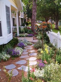 a garden with various flowers and plants in front of a white picket fenced house